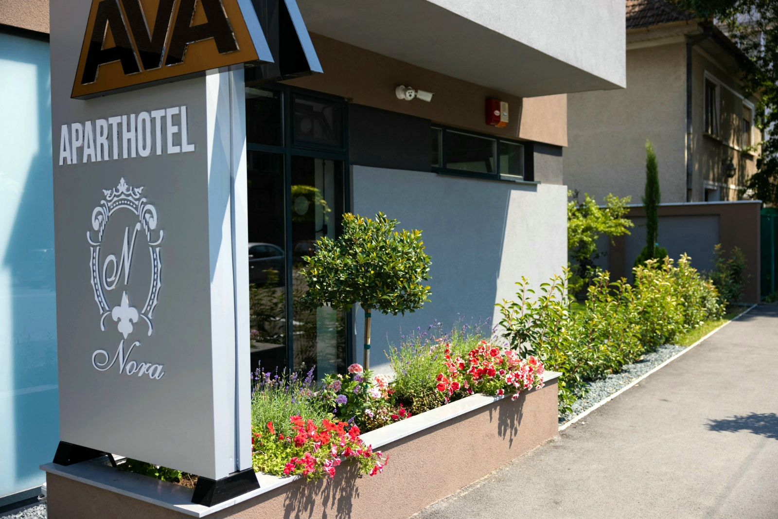 An apartment hotel with a sign reading 'APARTHOTEL Nora' and a small garden with various plants and flowers.