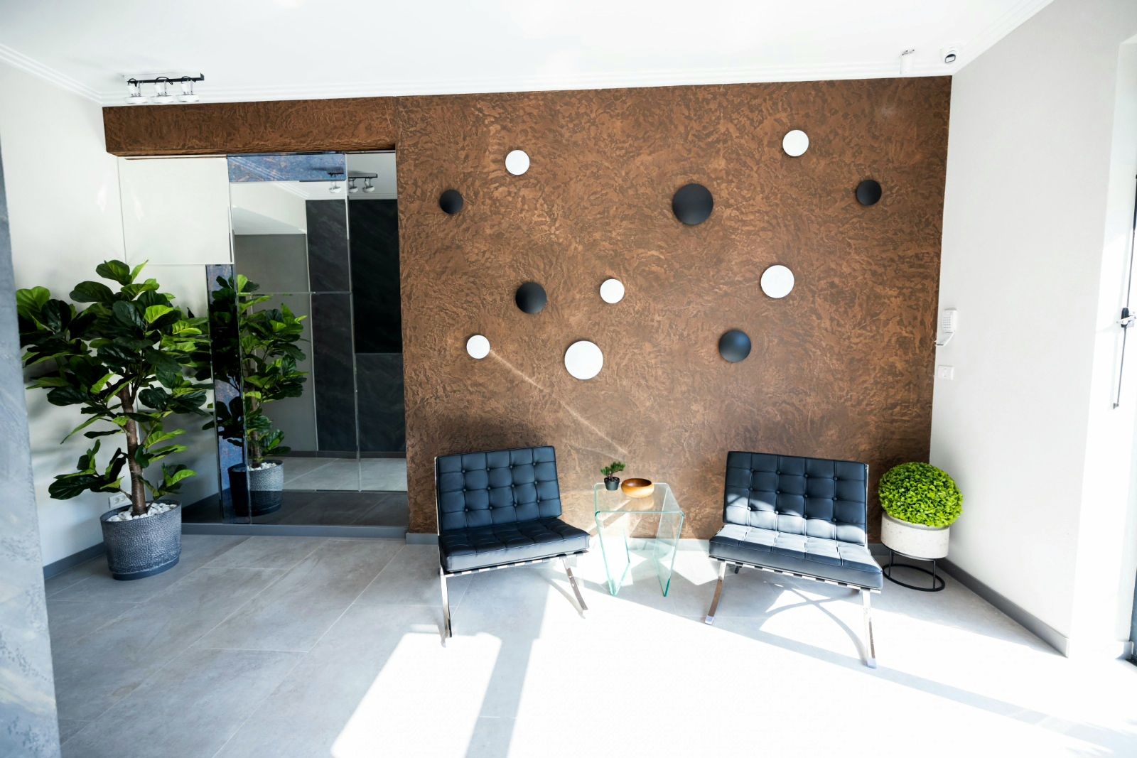 A modern reception area with two black upholstered chairs, a glass coffee table, and a decorative wall with circular cutouts.