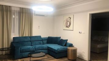 A living room with a blue sectional sofa, a round coffee table, and a small side table. There is a painting on the wall and a door leading to another room.