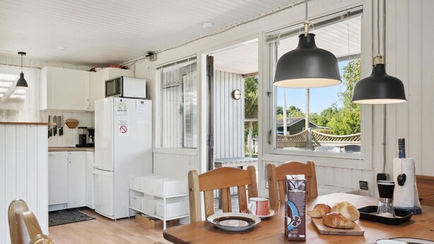 Een keuken met witte kastjes, een magnetron en een koelkast. Er is een eettafel met stoelen, een koffiezetapparaat en een schaal met croissants.