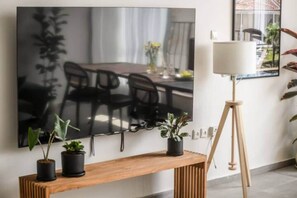 A flat-screen TV mounted on a wall, a wooden console table with potted plants, and a floor lamp. - Spacious & Bright 3BR Apartment by SeaNRent (Tel Aviv-Jaffa)