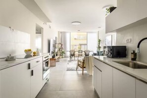 A modern kitchen with white cabinetry, a dining area with a table and chairs, and a view of a balcony. - Spacious & Bright 3BR Apartment by SeaNRent (Tel Aviv-Jaffa)