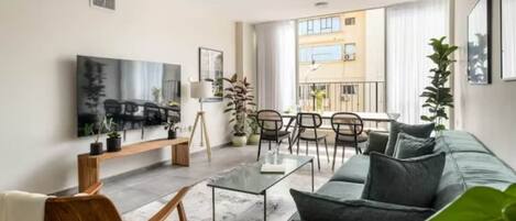 A modern living room with a sofa, coffee table, and chairs. There is a flat-screen TV mounted on the wall and a balcony with a railing.
