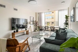 A modern living room with a sofa, coffee table, and chairs. There is a flat-screen TV mounted on the wall and a balcony with a railing. - Spacious & Bright 3BR Apartment (Tel Aviv-Jaffa)
