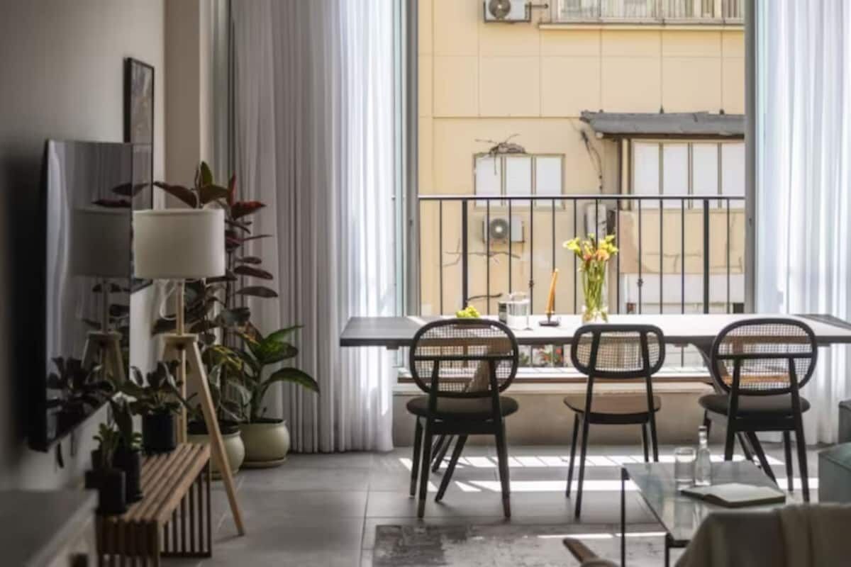 Un salon moderne avec balcon, comprenant une table à manger avec des chaises, des plantes en pot et une grande fenêtre donnant sur un immeuble.
