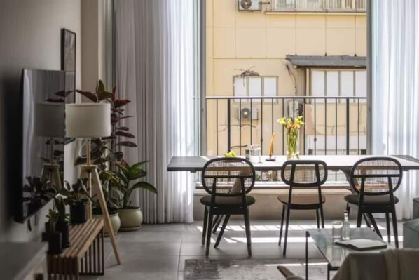 A modern living room with a balcony, featuring a dining table with chairs, potted plants, and a large window with a view of a building.