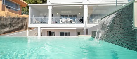 Un edificio de dos pisos con piscina, cascada y balcón con mobiliario exterior.