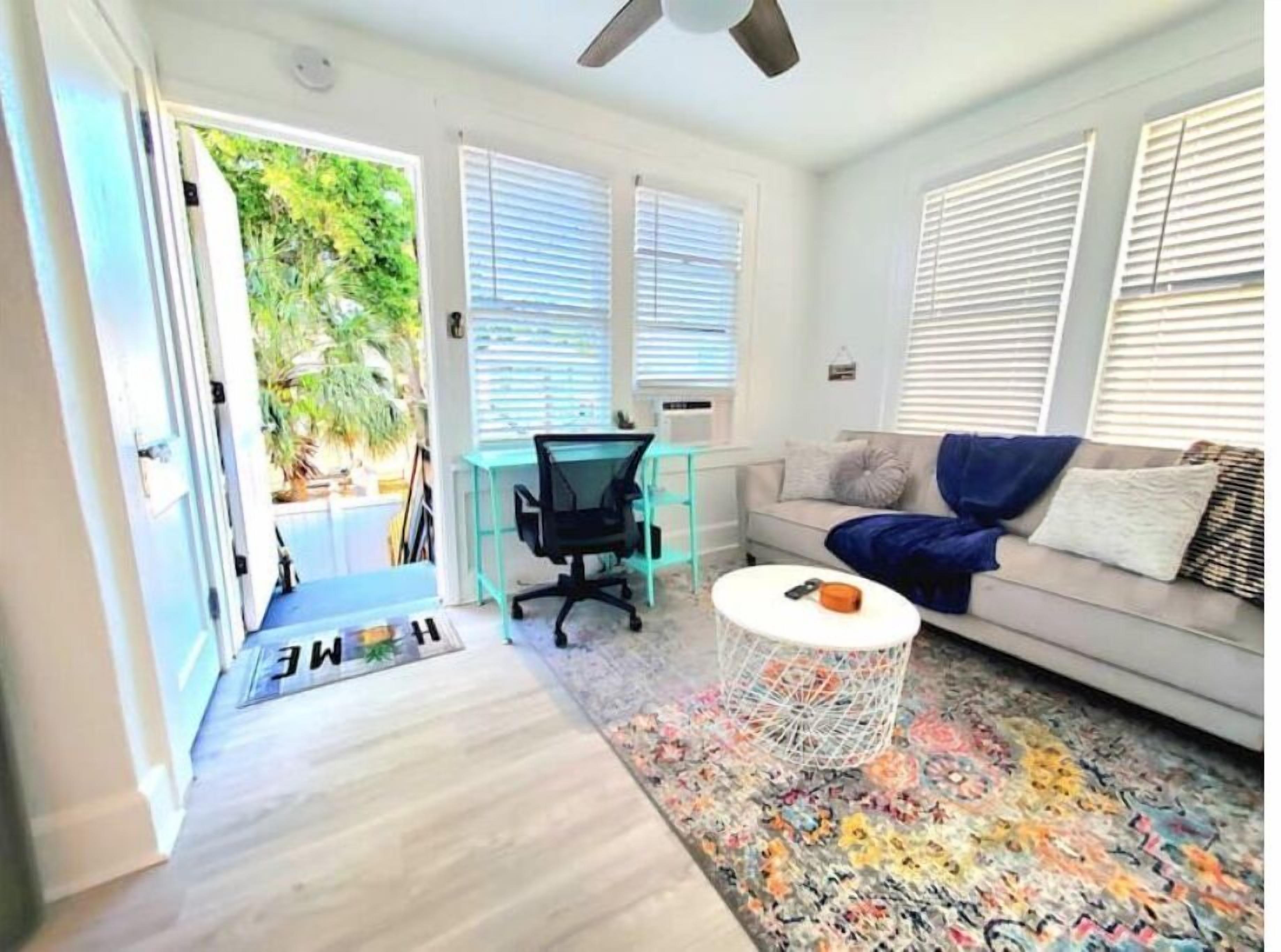 A modern living room with a sofa, a coffee table, and a desk with a chair. There is a colorful rug on the floor and a door leading outside.