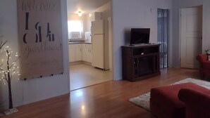 Un salon moderne avec un plancher en bois, un canapé rouge, un téléviseur posé sur un meuble en bois, et une cuisine visible à travers une porte ouverte.