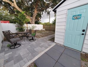 Une terrasse avec une table et des chaises, une porte bleue et, au fond, une clôture blanche.