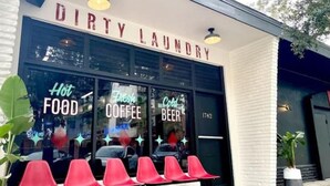 Une devanture de magasin avec une enseigne indiquant « DIRTY LAUNDRY » et des néons publicitaires pour « NOURRITURE CHAUDE », « CAFÉ FRAIS » et « BIÈRE FROIDE ».