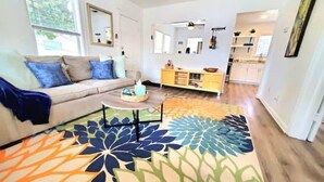 Un salon avec un canapé, une table basse et un tapis coloré. On aperçoit à l’arrière-plan un coin cuisine avec des placards et un plan de travail.