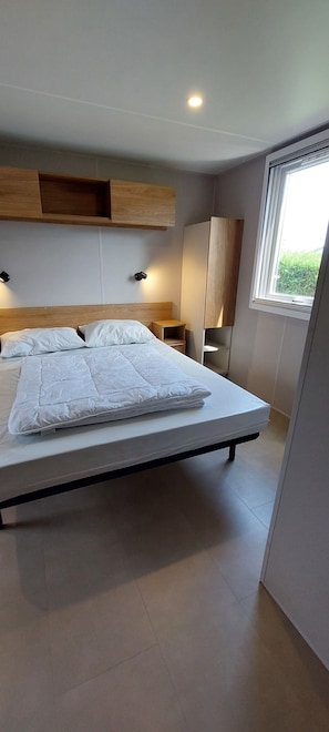 Een moderne slaapkamer met een groot bed, ingebouwde houten planken erboven, een nachtkastje en een raam met uitzicht op groen.