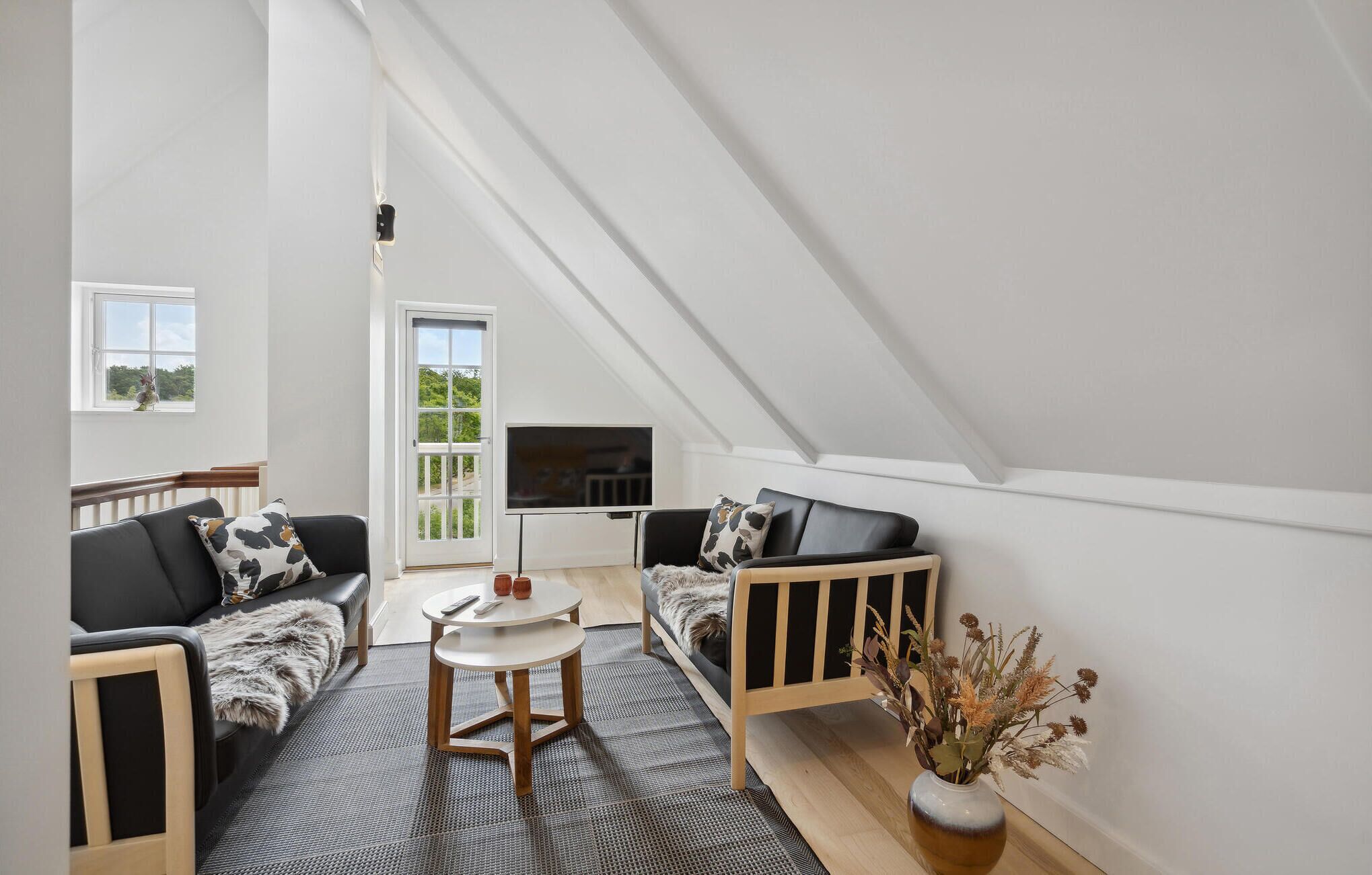 Een moderne loft slaapkamer met een schuine kap, een groot raam, een zwarte leren bank, een houten bankje en een kleine ronde tafel.