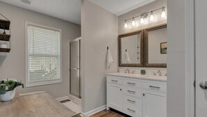 A bathroom with a shower, a sink with a mirror, and a window with blinds.
