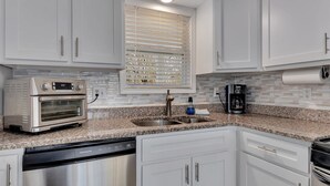 A modern kitchen with white cabinets, a built-in oven, a dishwasher, and a window with blinds.