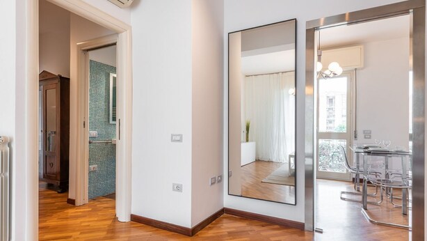 Un apartament modern amb sòl de fusta, un mirall gran i una zona de dinar amb cadires transparents.