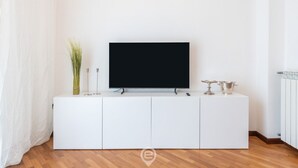Un saló modern amb un televisor de pantalla plana sobre un moble blanc, un vas amb plantes verdes i una taula petita amb coberts.