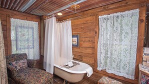 Interno rustico di una baita con pavimento in legno, una vasca da bagno vintage, un divano decorato e tende in pizzo.