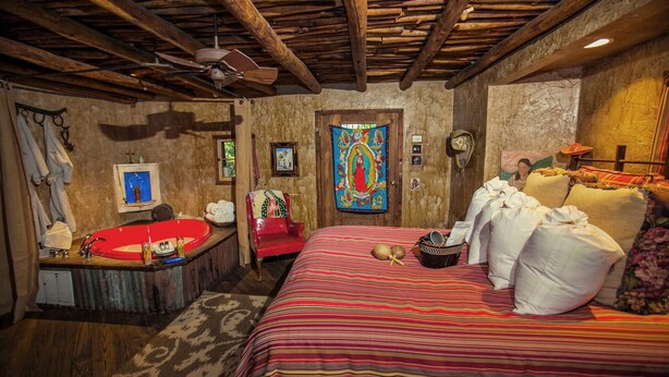 Een landelijke kamer met een houten plafond, een rode badkuip, een bed met een kleurrijke gestreepte dekbedovertrek en een religieuze schildering aan de muur.