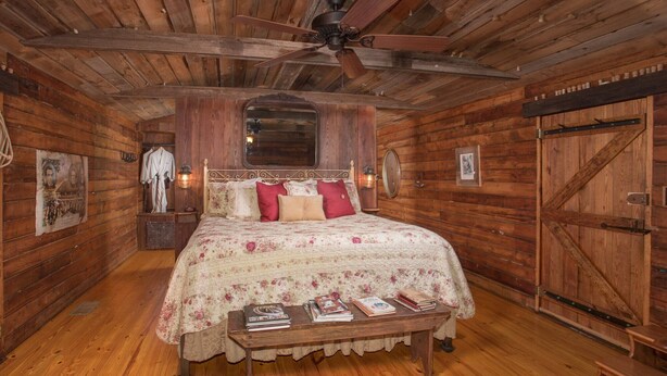 A wooden cabin bedroom with a bed, a ceiling fan, a wooden door, and a small table with books. - 7F Lodge - Batts Ferry Cabin w/Breakfast - Log Cabin w/ Jacuzzi Tub and Fire Pit (College Station)
