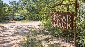 Een bord met de tekst 'SUGAR SHACK' in een bebost gebied, met een oldtimer-truck op de achtergrond.