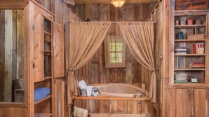 Eine rustikale Holzhütte mit einer Holzbadewanne, einer kleinen Bücherablage und einem Fenster mit Vorhängen.