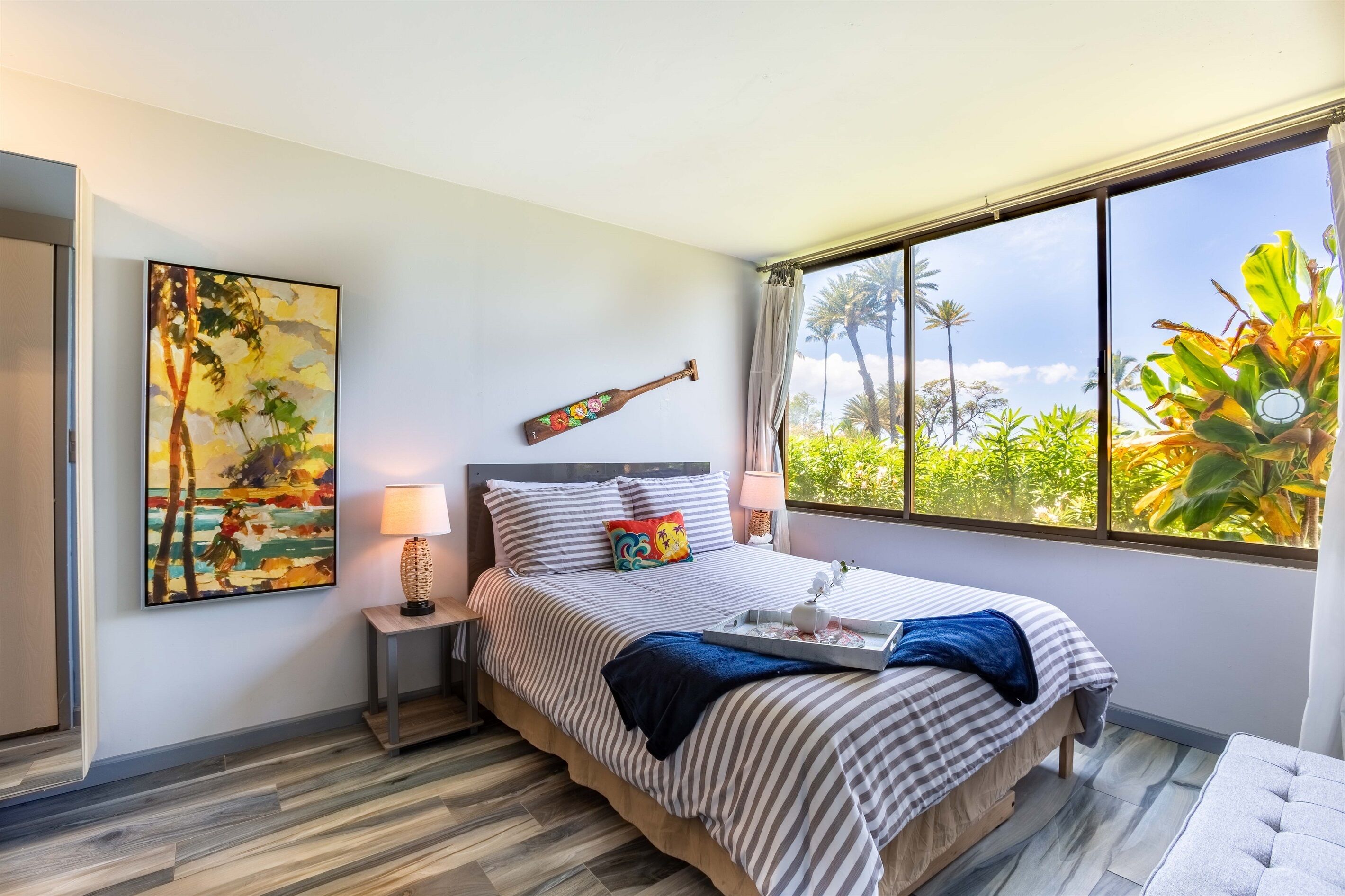 Un dormitorio con cama, mesita de noche, lámpara, un cuadro y ventana con vista a zonas verdes.