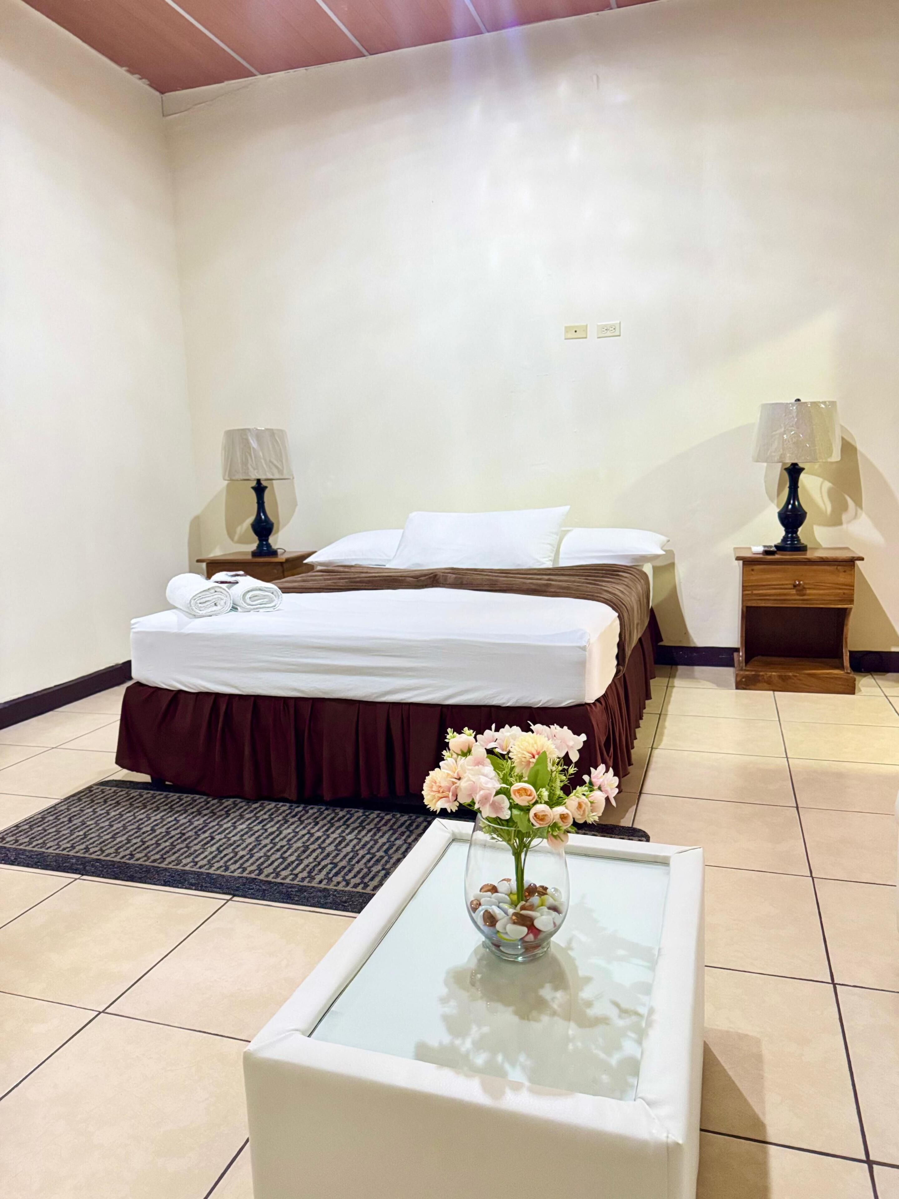 A hotel room with a bed, two bedside tables with lamps, a small table with a vase of flowers, and a rug.