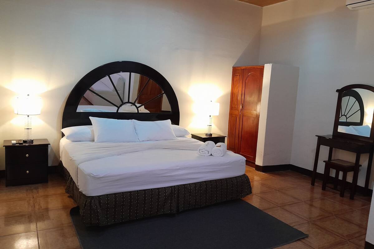 Una cama bien tendida con sábanas blancas, dos lámparas de noche, un armario de madera y una silla con espejo.