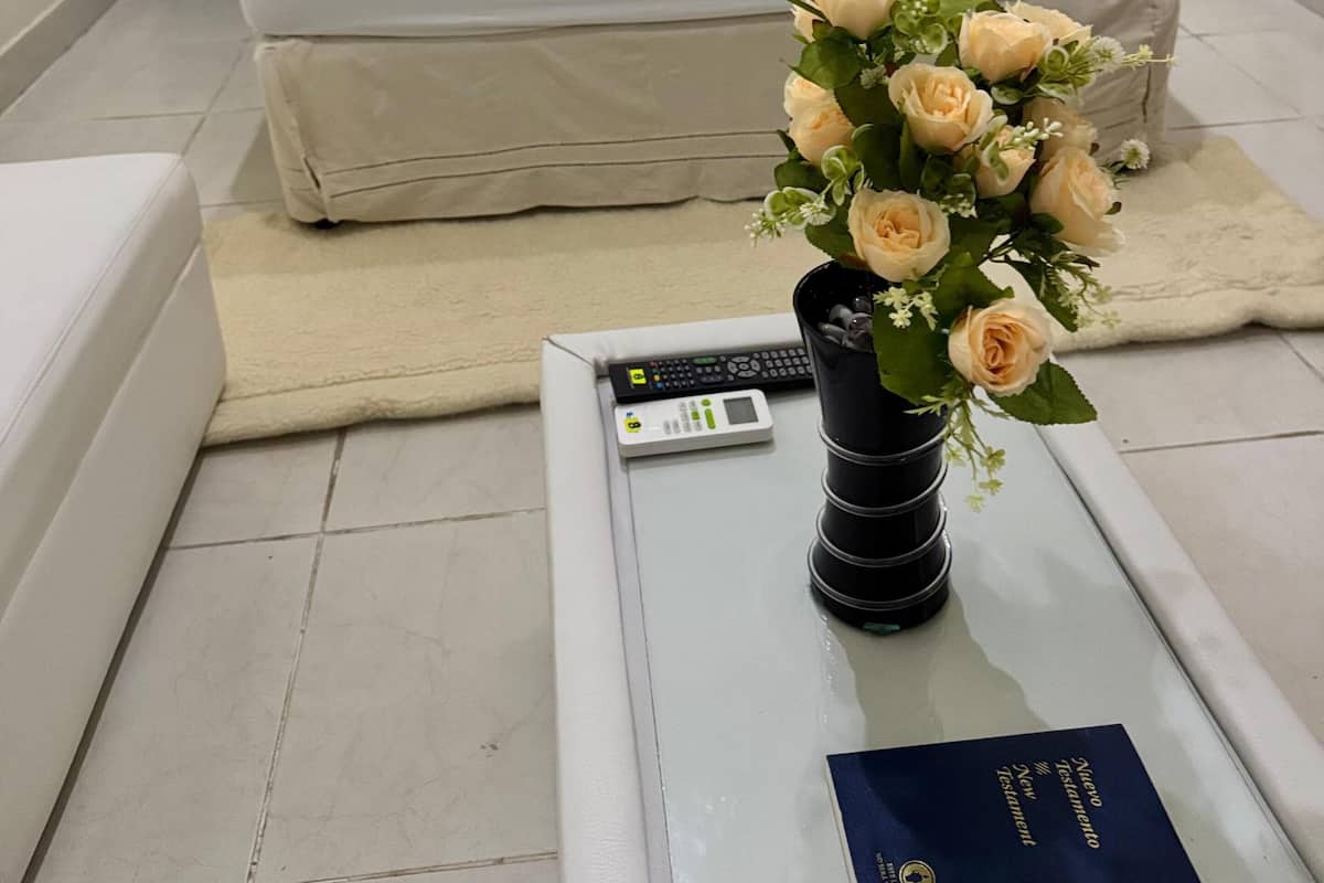 Habitación de hotel con cama, mesita de noche con control remoto, jarra con flores y un libro sobre la mesa.