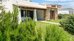 A white building with a stone entrance, a covered patio with a table and chairs, and a well-maintained garden.