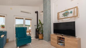 Een woonkamer met een smaragdgroene bank, een houten tv-meubel met een flatscreen-televisie, een grote potplant en uitzicht op de buitenlucht door een schuifdeur.