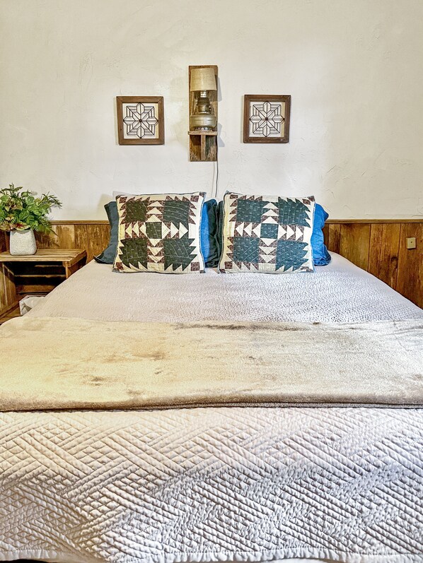 Una cama con colcha acolchada, dos almohadones decorativos, una mesita de noche con una planta y dos lámparas fijadas a la pared.