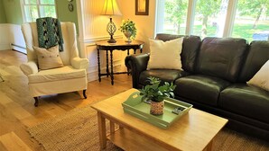 A living room with a wooden coffee table, a white armchair, a black leather sofa, and a small side table with a lamp. - Luxury Country Home, Spacious and Cozy, with Sun Room (Marionville)