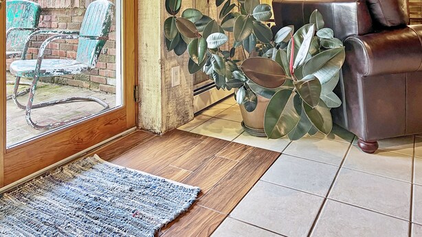 A living room with a brown leather sofa, a large potted plant, and a view through a glass door to an outdoor area with a brick wall and a blue chair. - Luxury Country Home, Spacious and Cozy, with Sun Room (Marionville)