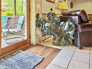 Una sala de estar con un sofá de cuero marrón, una planta grande en maceta y vistas a un espacio exterior con un muro de ladrillo y una silla azul.