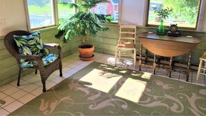 A sunlit room with a large window, a potted plant, a wicker chair, a wooden table, and a chair with a cushion. - Luxury Country Home, Spacious and Cozy, with Sun Room (Marionville)