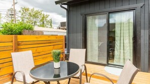 Un patio al aire libre con una mesa redonda y sillas, una valla de madera, y un edificio con fachada oscura y grandes puertas correderas de vidrio.