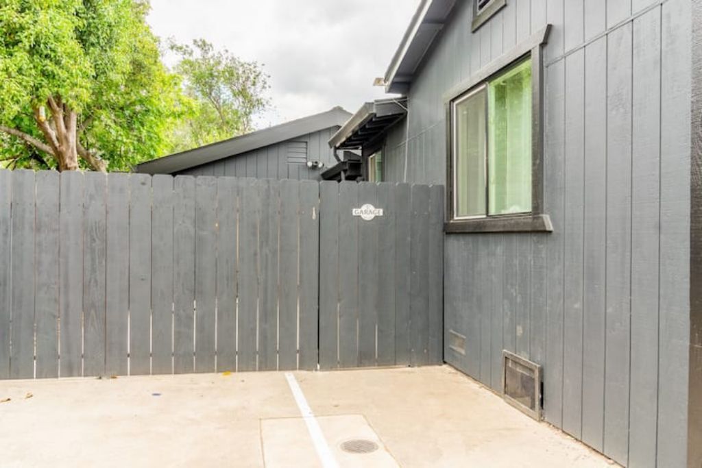 A grey wooden fence with a garage sign, a concrete patio, and a window with a green curtain.
