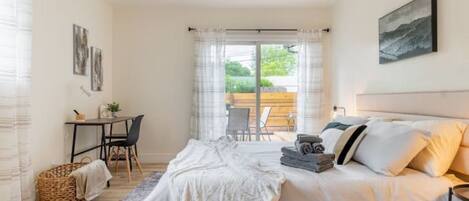 Un dormitorio con cama, escritorio y silla. Hay una ventana con cortinas, un cuadro en la pared y vistas al exterior.