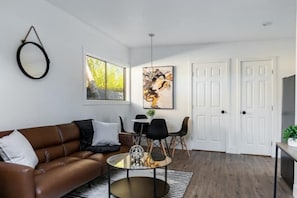 Uma sala de estar moderna com um sofá de couro marrom, uma mesa de centro de vidro e um espaço de jantar com uma mesa branca e cadeiras pretas. Há uma grande pintura abstrata na parede e uma porta branca ao fundo.