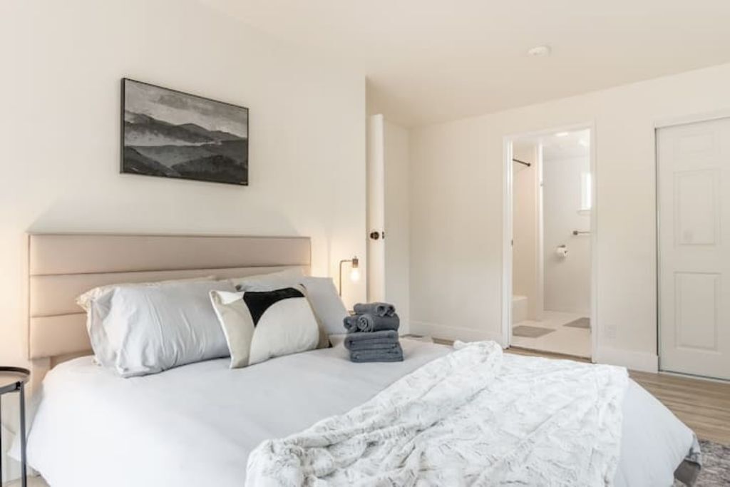 A modern bedroom with a large bed, a headboard, a bedside table, a nightstand, a wall-mounted light, a framed picture, and a door leading to a bathroom.