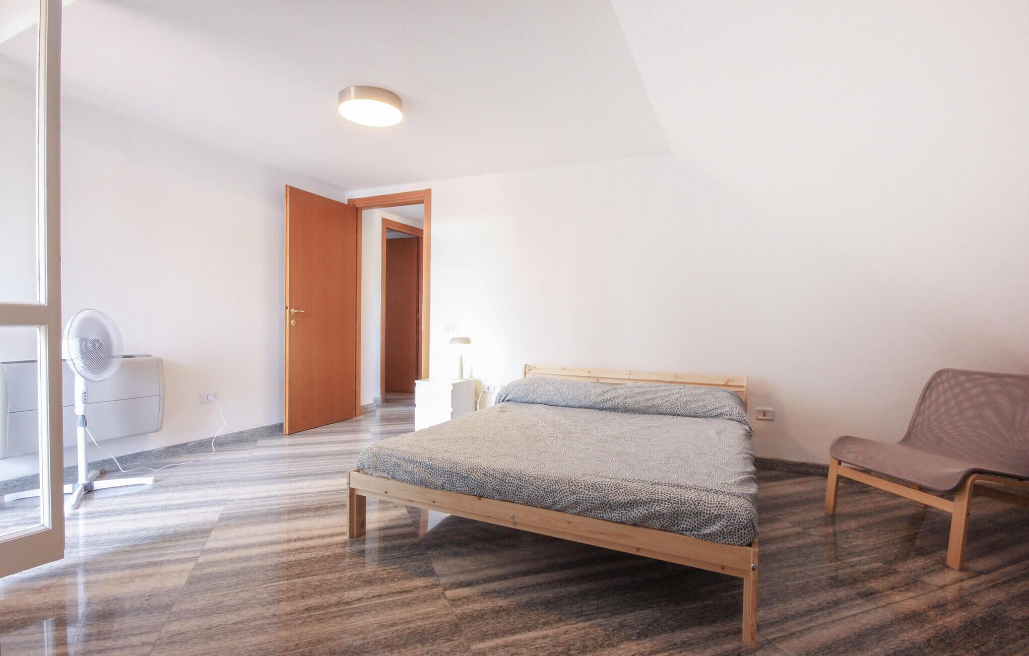 A bedroom with a bed, a chair, a desk, and a fan.