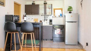 Cocina compacta con microondas, estufa y refrigerador. Hay dos taburetes negros y una pequeña planta en maceta sobre la encimera.