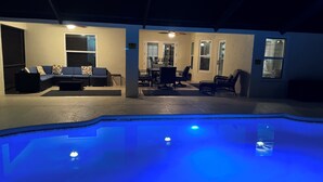 A pool area with a blue-lit pool, a sofa, and a dining table with chairs. - Amazing and spacious home. 4be2Bth, and beautiful screened private pool. (Port St. Lucie)