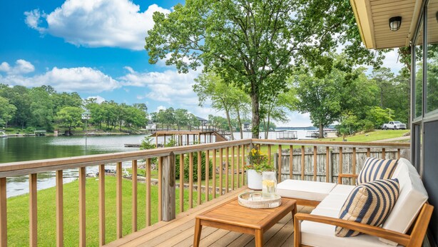 Una terraza de madera con vistas a un lago, un sofá blanco con almohadones a rayas, y una mesita con una bandeja y una jarrera.