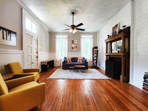 Una sala de estar con piso de madera, un sofá, un sillón amarillo, un ventilador de techo y una chimenea.