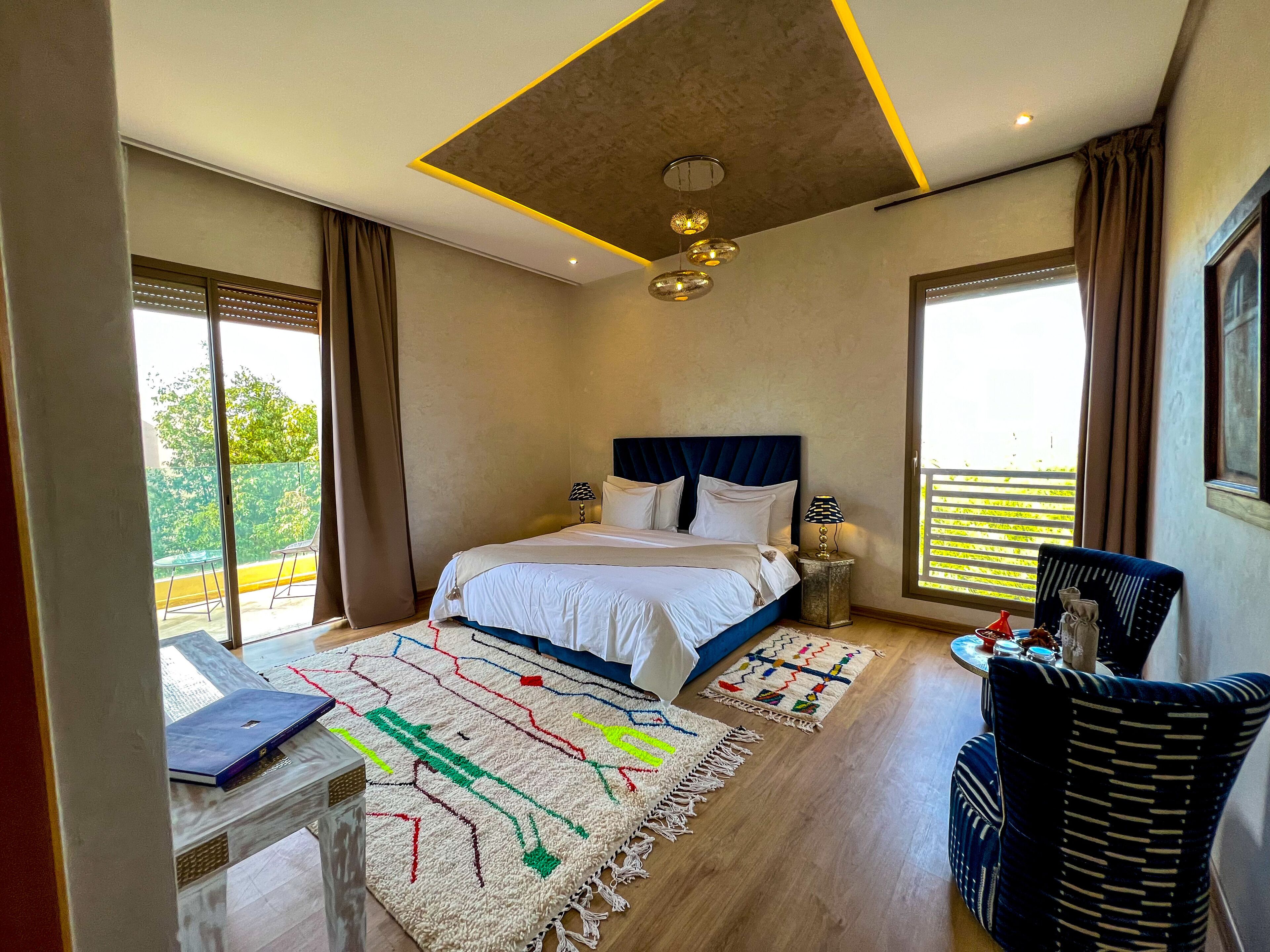A modern bedroom with a large bed, a desk, and a chair. There are two windows and a balcony visible.