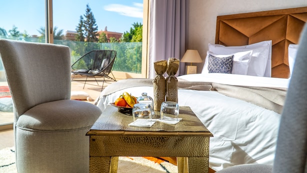 A hotel room with a bed, a chair, a table with fruits and glasses, and a view of trees and buildings. - Riad villa saphir & SPA (Marrakech)